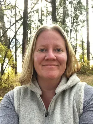 Person smiling in a gray hoodie in a sunlit forest with tall trees.