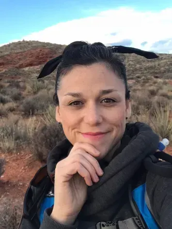 Person smiling outdoors with a backpack, standing in a rocky desert landscape under a blue sky
