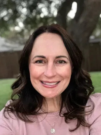 Smiling woman with dark hair outdoors, wearing a light pink top and necklace.