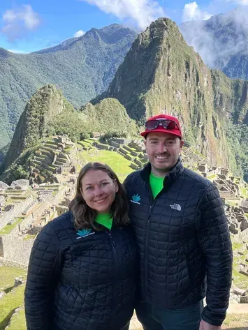 Two people in black jackets posing at Machu Picchu with mountains and stone terraces behind them