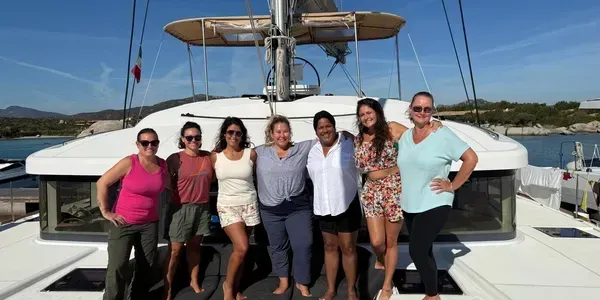 Six people smiling on a boat deck at the marina, with calm water and blue sky behind them.