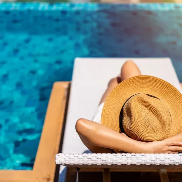 Person reclining on a poolside lounge chair beside bright blue water, wearing a tan sunhat.
