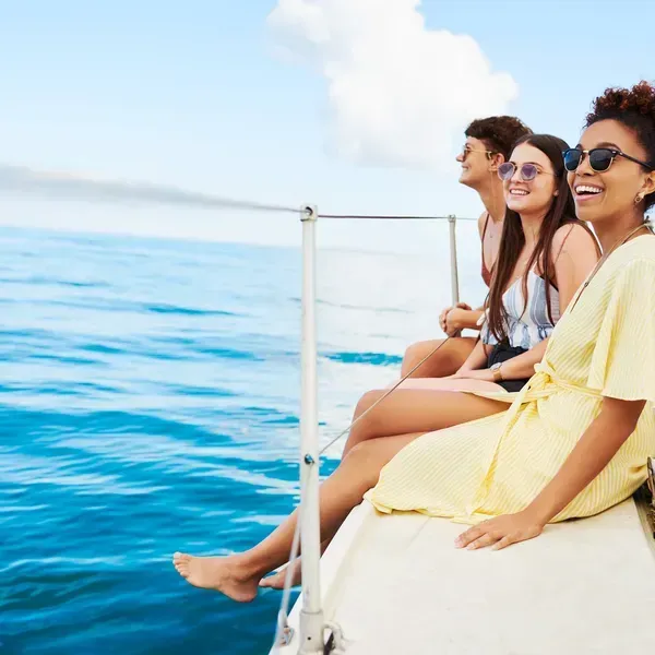 Three people sit on a boat deck above blue ocean, smiling in sunglasses.
