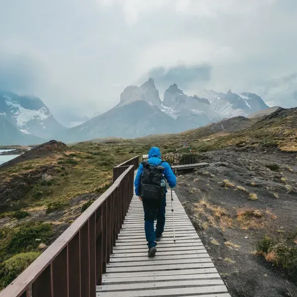 Person in a blue jacket walking on a wooden boardwalk through a rocky mountain landscape.