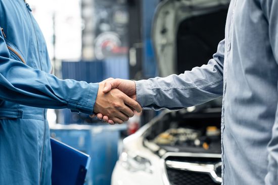 Two people shaking hands beside a car with its hood open in a workshop