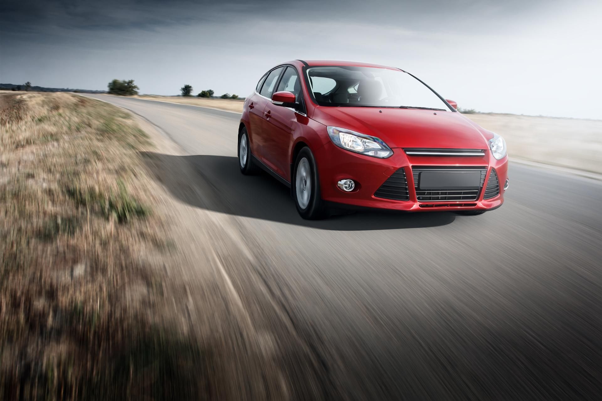 Red compact car driving on a winding road through a grassy hillside under a cloudy sky