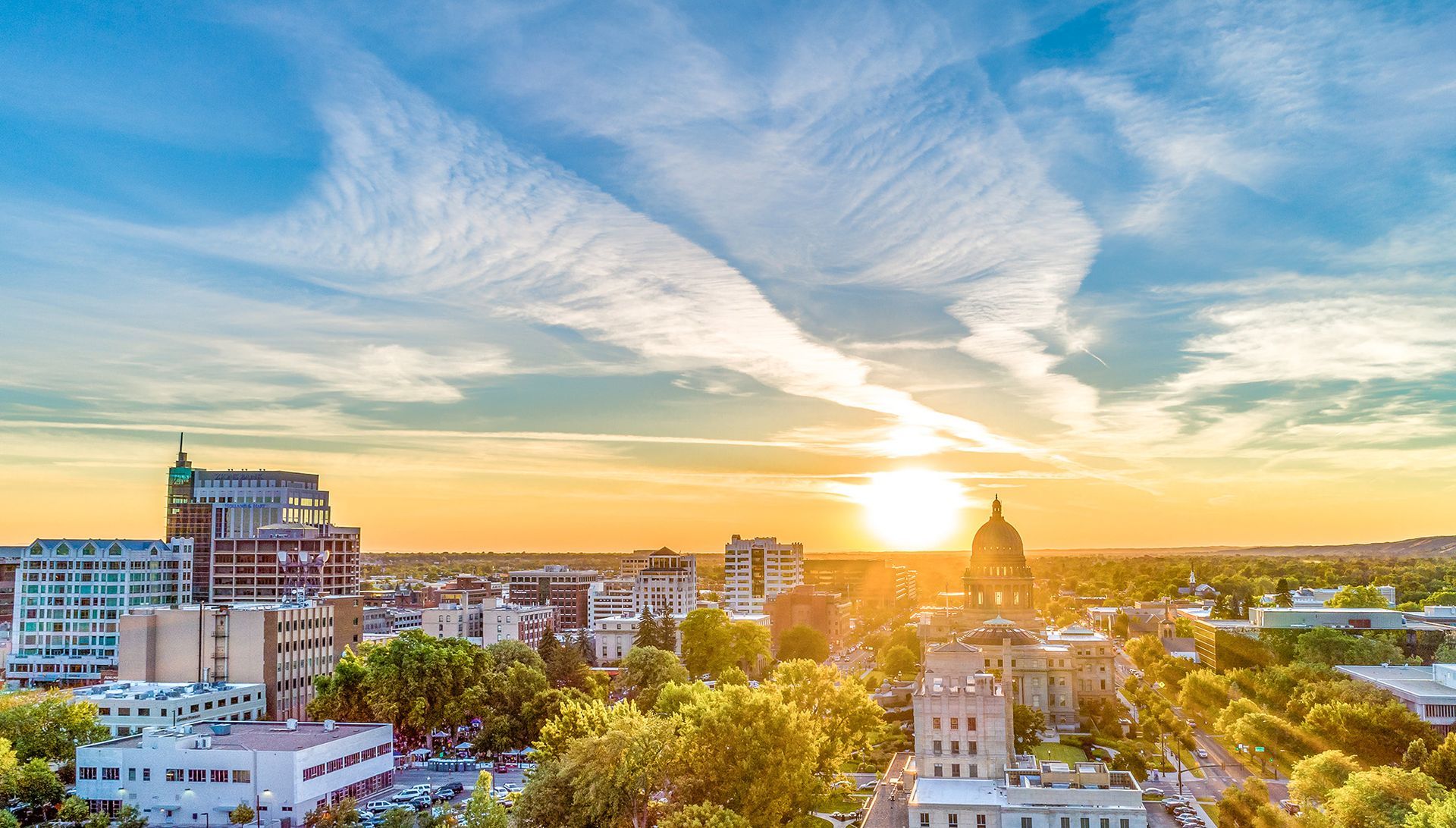 City skyline at sunset with golden light, scattered clouds, and trees in the foreground