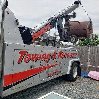 Tow truck lifting a rusty dumpster in a residential area. Truck is silver, with red accents and the name Fred's Towing.