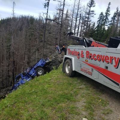A blue car is being towed from a hillside by a tow truck; near dead trees.
