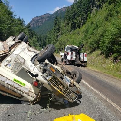 Overturned camper on roadside with tow truck, mountains in background.