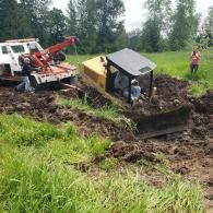 A bulldozer stuck in mud being pulled out by a tow truck on a grassy field.