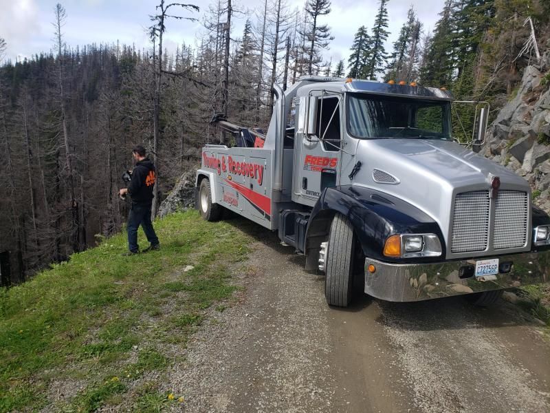 Tow truck on dirt road next to person, wooded hillside in background.