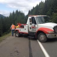 Tow truck on a road beside a forest; workers in vests, preparing to tow a vehicle.