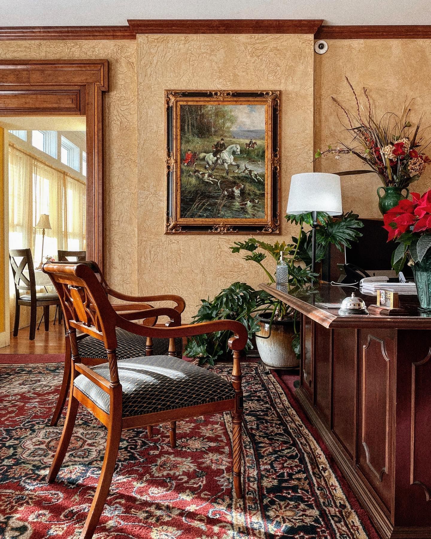 Antique desk and chairs in a room with a painting, plants, and patterned rug.