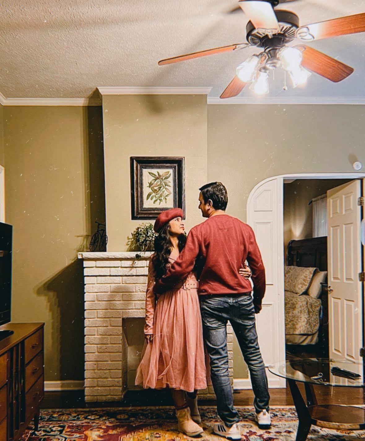 Couple in a living room, embracing. Woman wears pink dress and hat, man in red sweater. Fireplace, art, and open doorway.