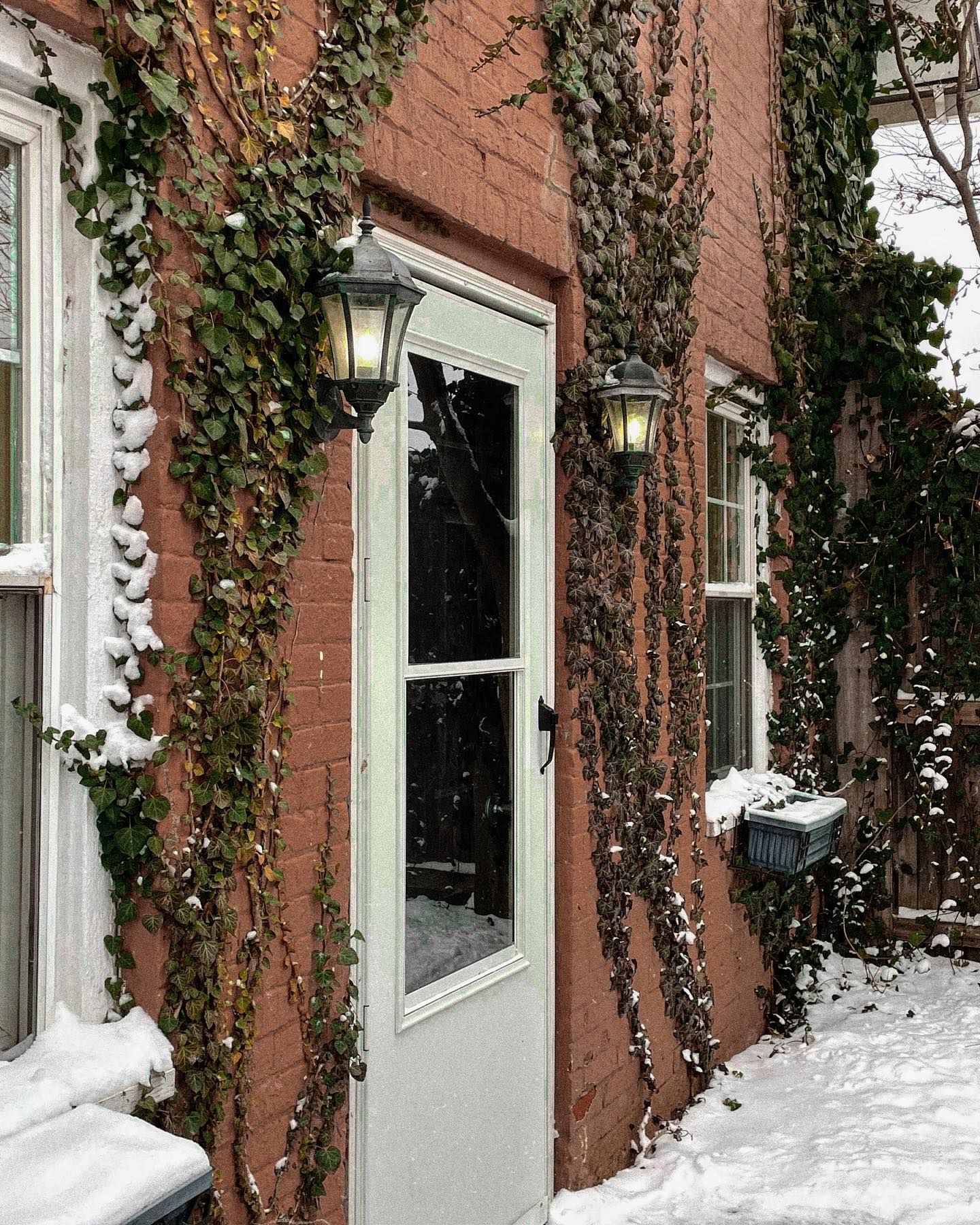 Brick building with door, windows, and vines. Snow covers the ground. Two lanterns flank the door.