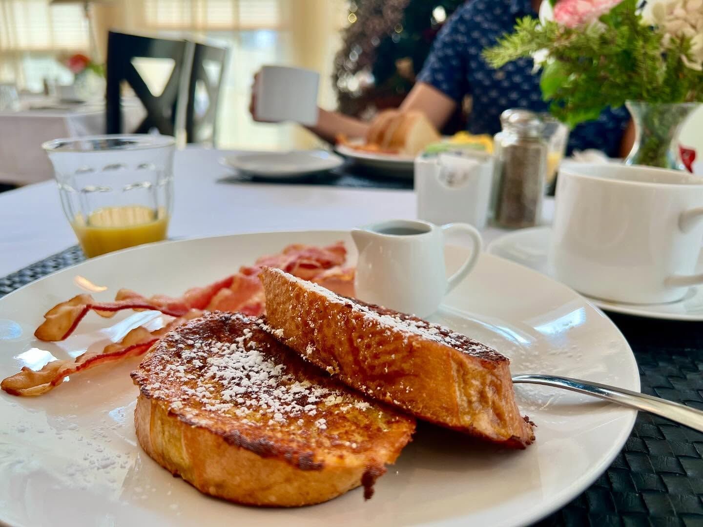 Breakfast plate with French toast, bacon, and syrup. A person is holding a coffee cup at a table.