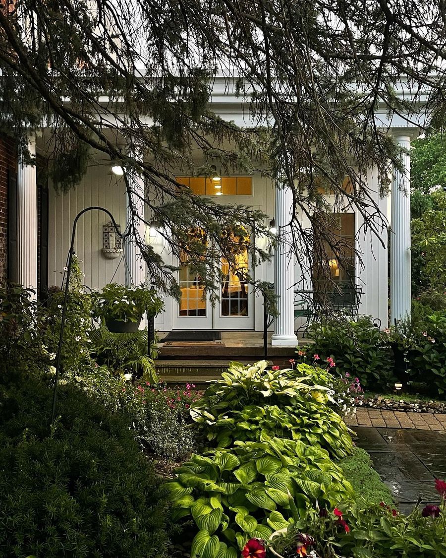 White-columned building with a porch, framed by lush greenery and a walkway.