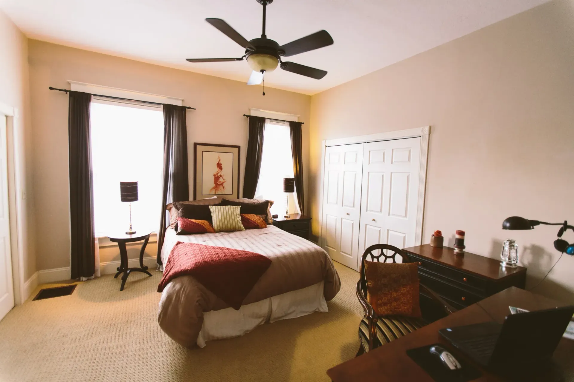Bedroom with bed, desk, closet, and ceiling fan. Brown, beige, and white colors. Sunlight through windows.