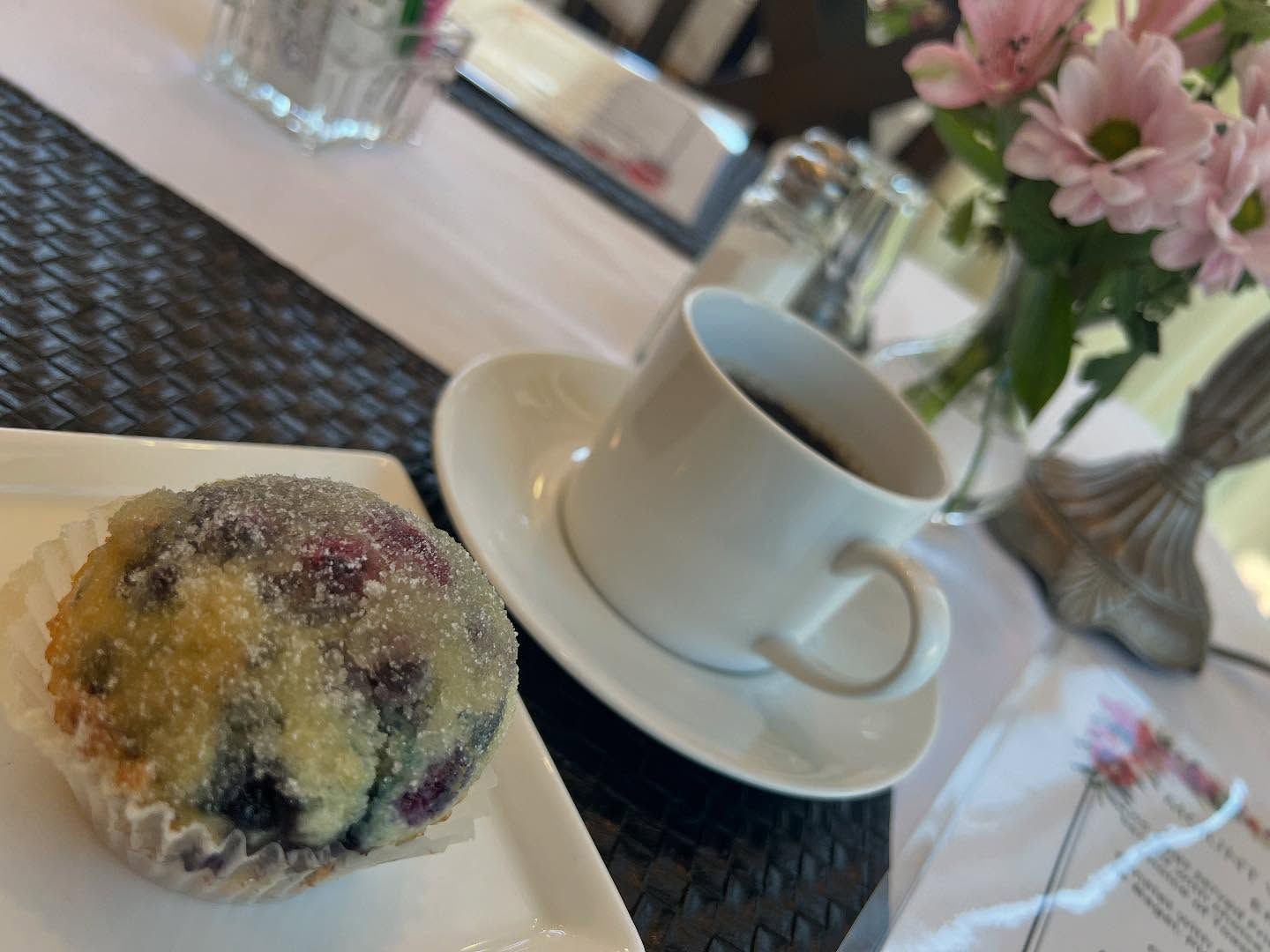 Muffin with sugar crystals, coffee cup on saucer, and pink flowers on a table.