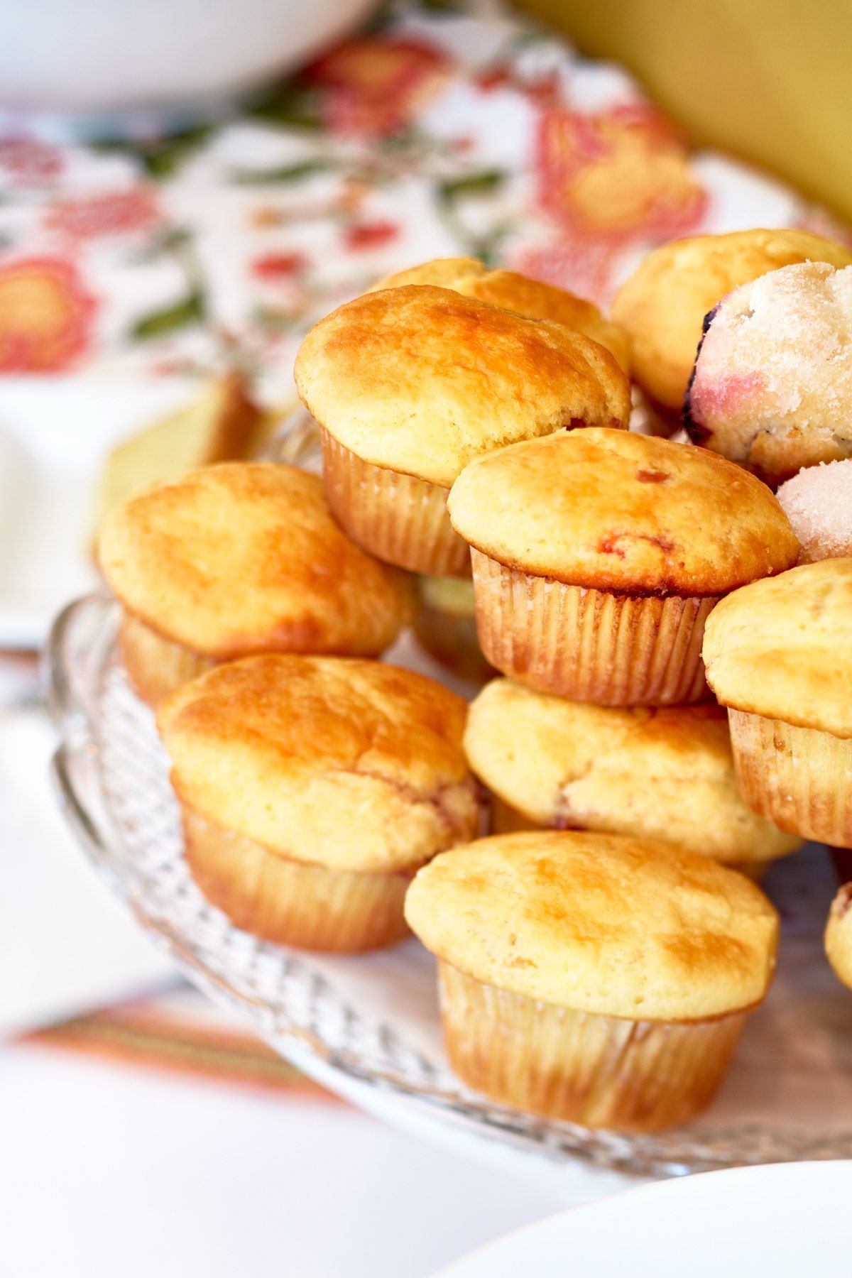 Pile of golden muffins on a glass serving plate, with a floral tablecloth in the background.