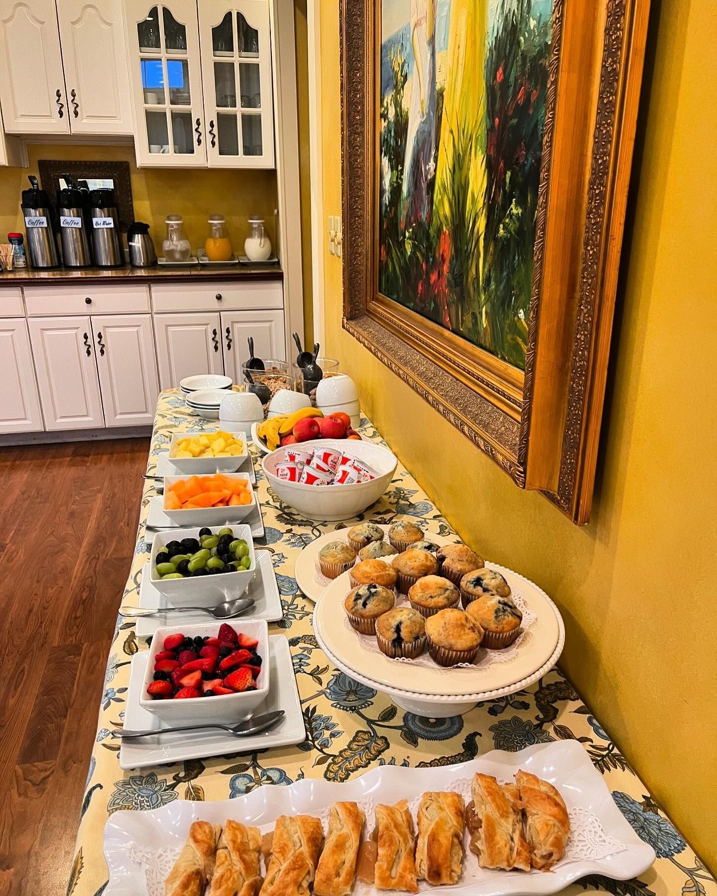 Breakfast buffet: muffins, pastries, fruit, and yogurt on a table next to a painting in a sunny room.