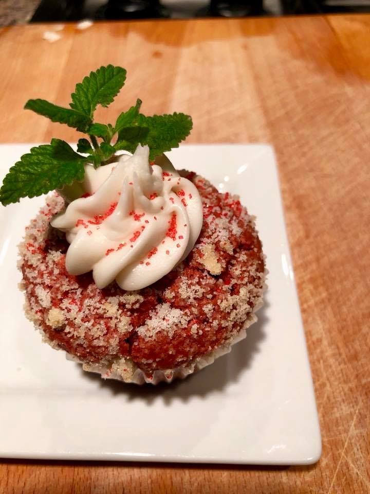 Red cupcake with white frosting, red sprinkles, and mint garnish on a white plate on a wooden surface.