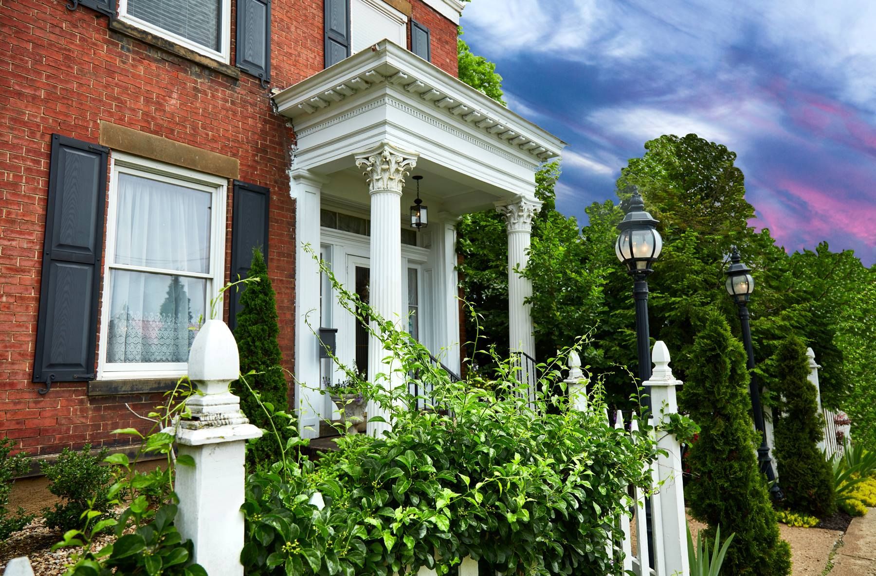 Red brick house with white porch, trim, and picket fence, green bushes, and a colorful sky.