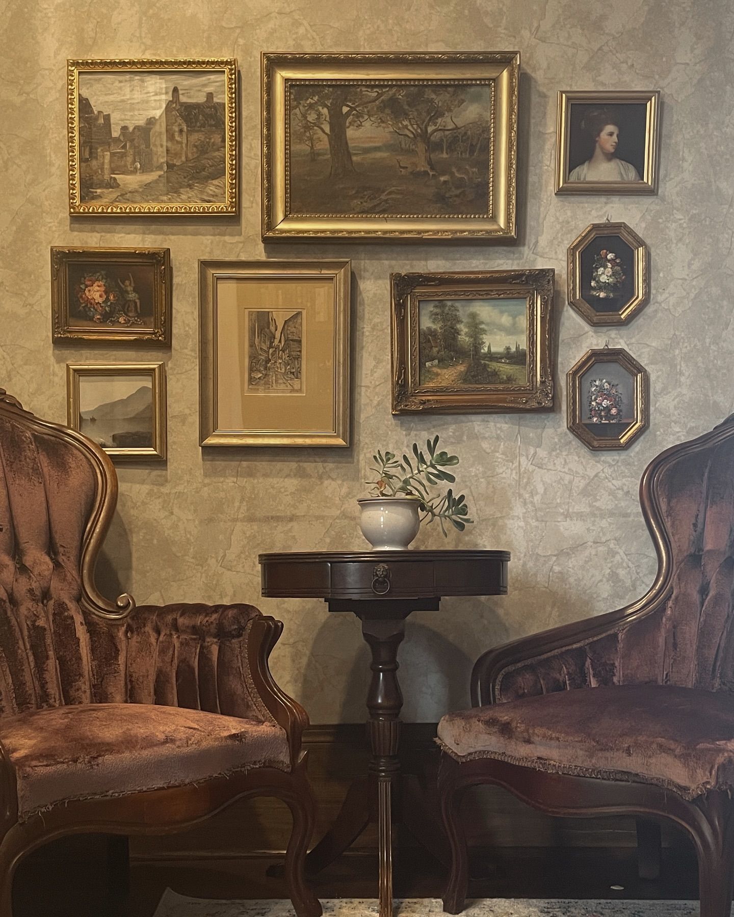 Two velvet chairs face a small table beneath a wall of framed vintage art.