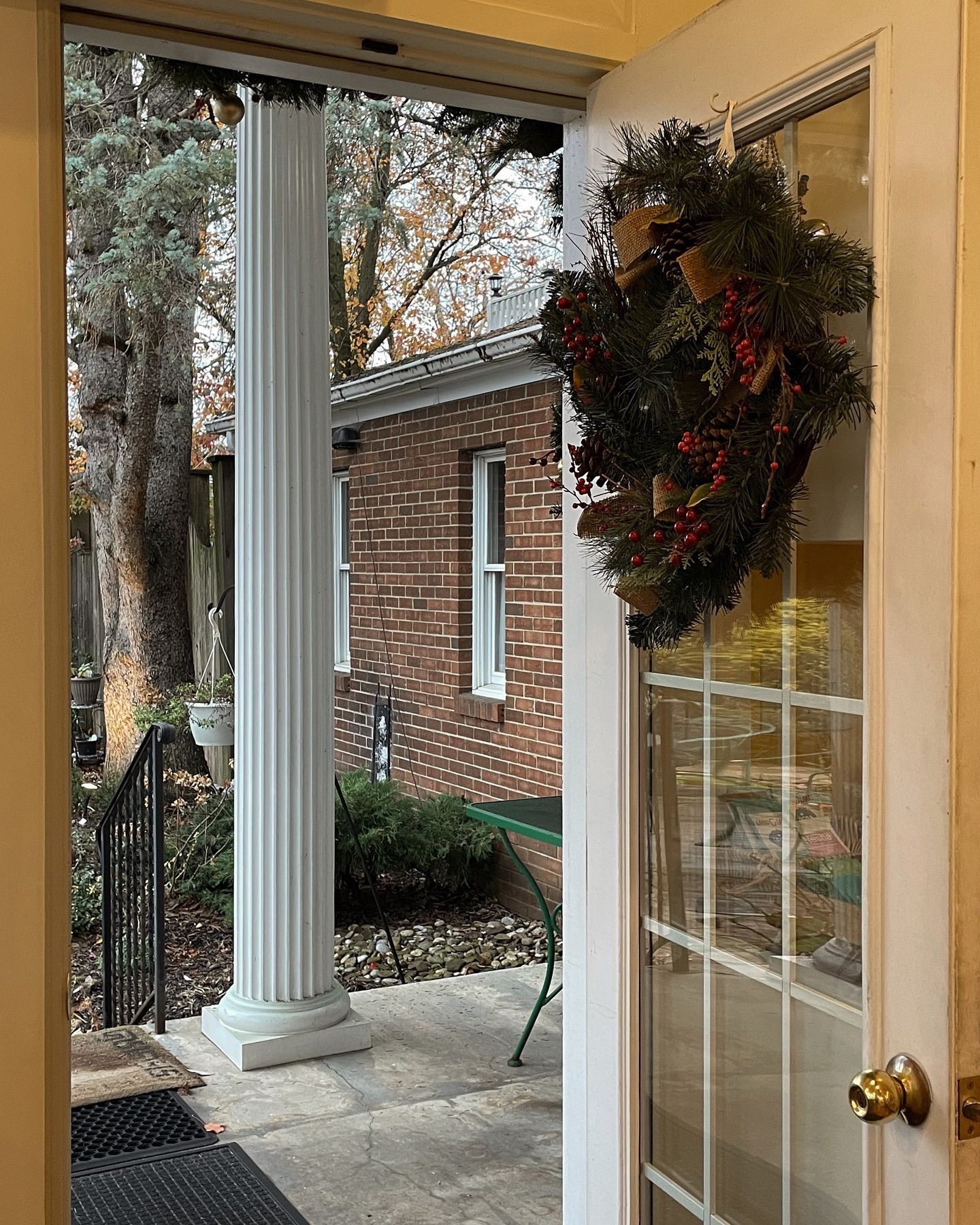 Open door with a Christmas wreath, leading to a porch with a column and a brick building in the background.