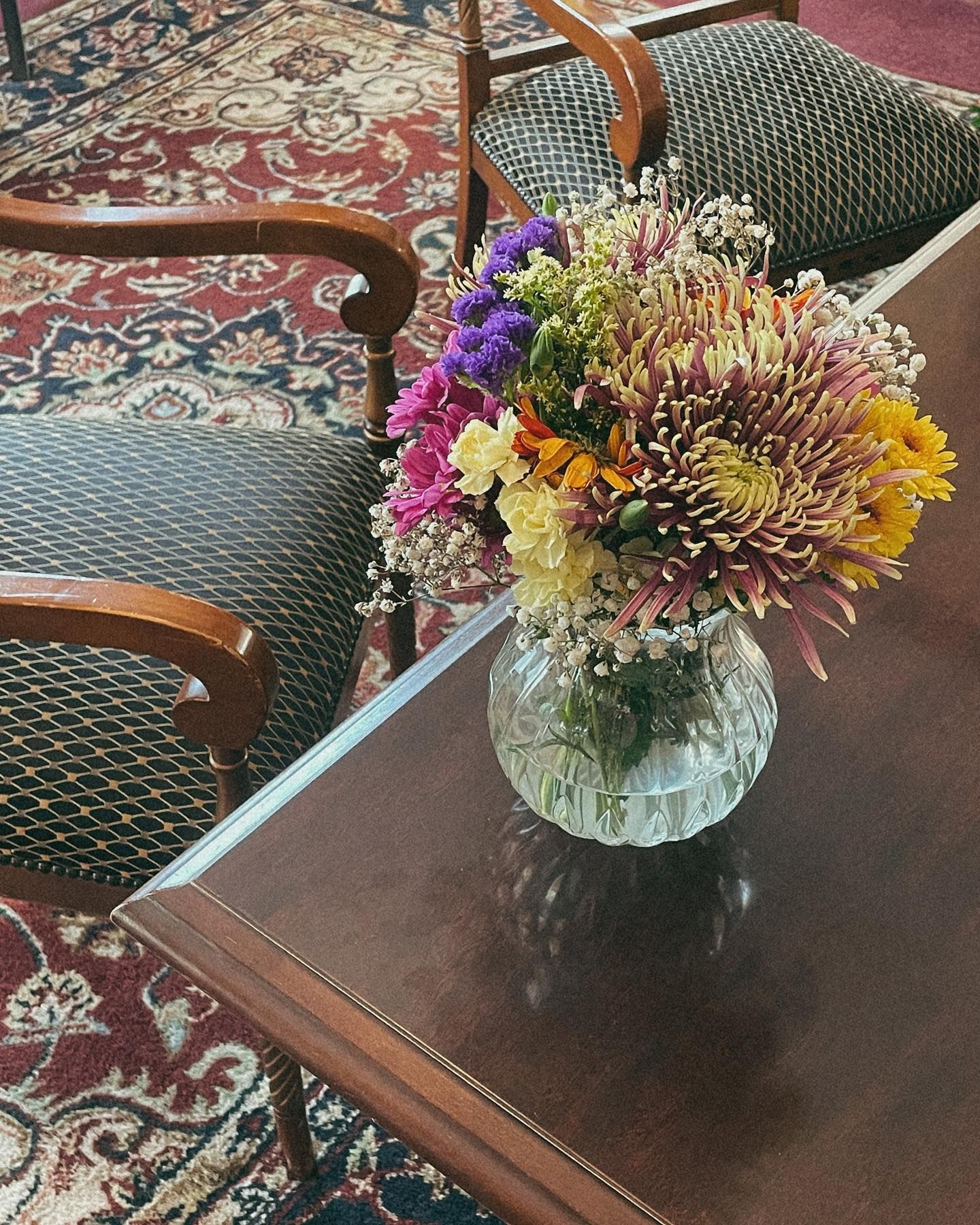 Colorful flowers in a glass vase on a brown table, chairs in the background, floral rug.