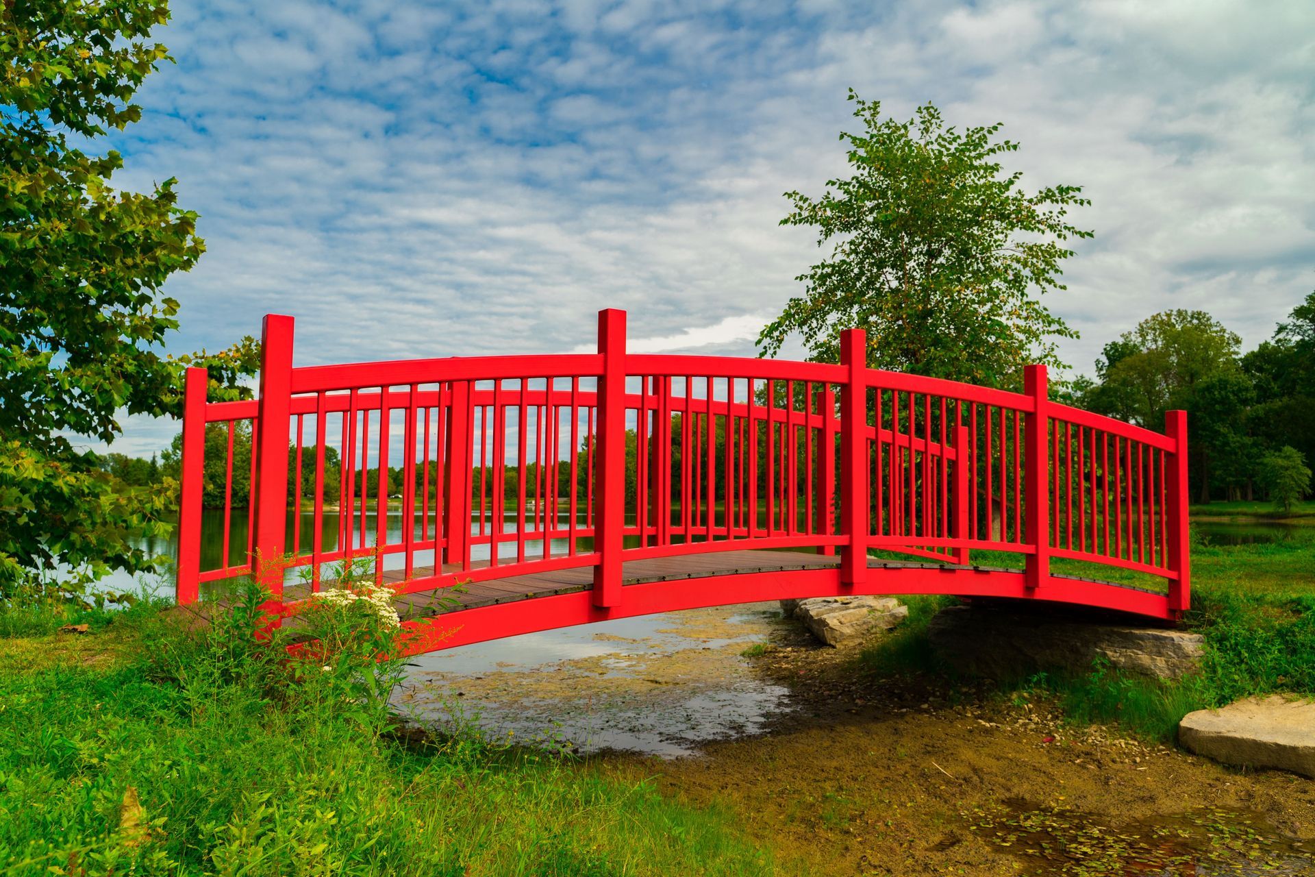 Bright red footbridge over a stream in a green park under a partly cloudy sky