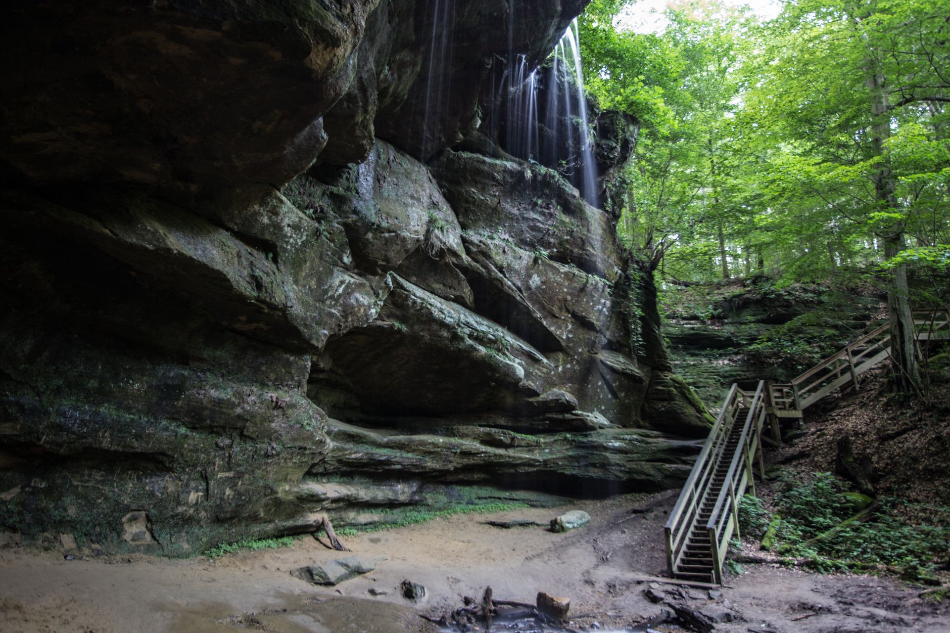 Rocky waterfall in a forest cave with a wooden staircase and mossy green surroundings