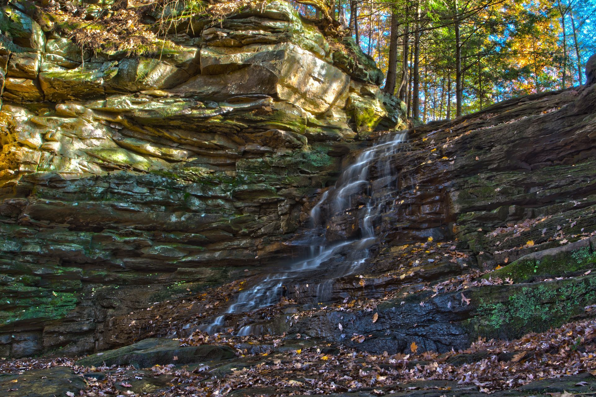 Small waterfall cascading over layered rock in a forest with autumn foliage.