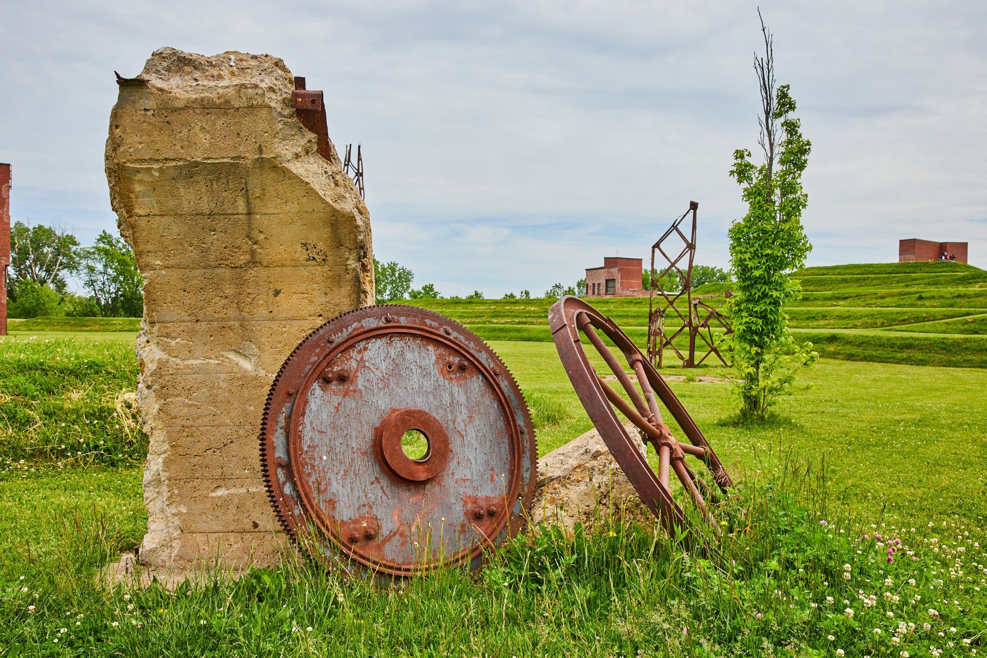 Rusty old wheel and metal frame beside a crumbling stone wall in a grassy field under a cloudy sky