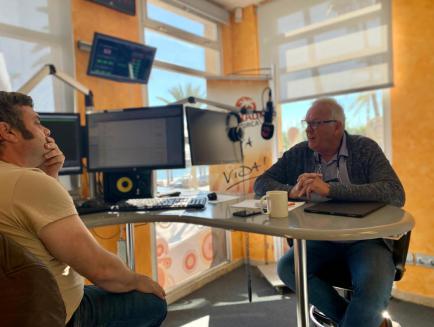 Dos hombres en una emisora ​​de radio; uno hablando por un micrófono, el otro escuchando. Escritorio, computadoras y ventana visibles.