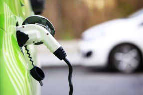 Vehículo eléctrico verde cargándose con el enchufe conectado. Coche blanco difuminado al fondo.
