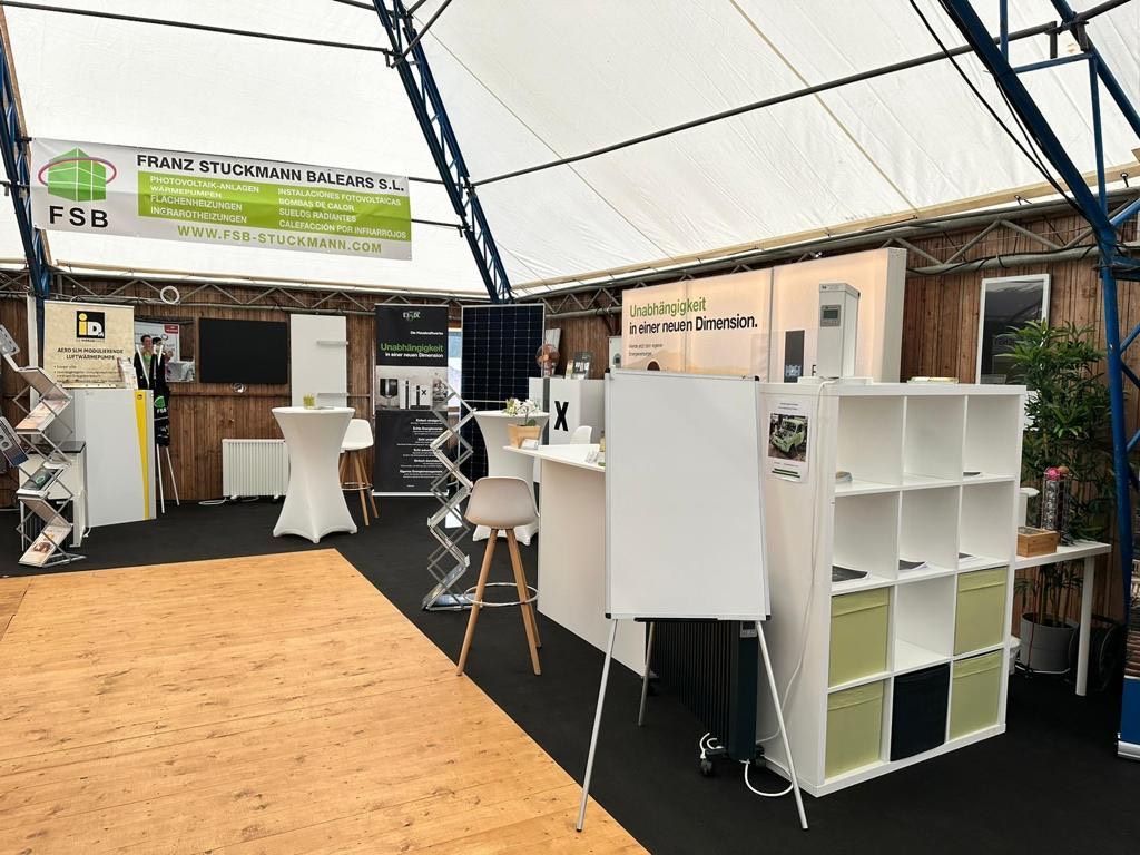 Stand de feria comercial en interior. Mesas de exposición, estanterías, pancartas, paneles solares, suelo de madera y techo de lona blanca.