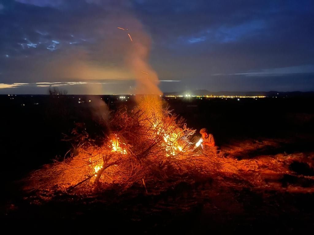 Gran hoguera nocturna con llamas anaranjadas y humo, contra un campo oscuro y luces de la ciudad.
