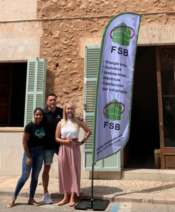Tres personas posan junto a una bandera del FSB frente a un edificio con persianas.