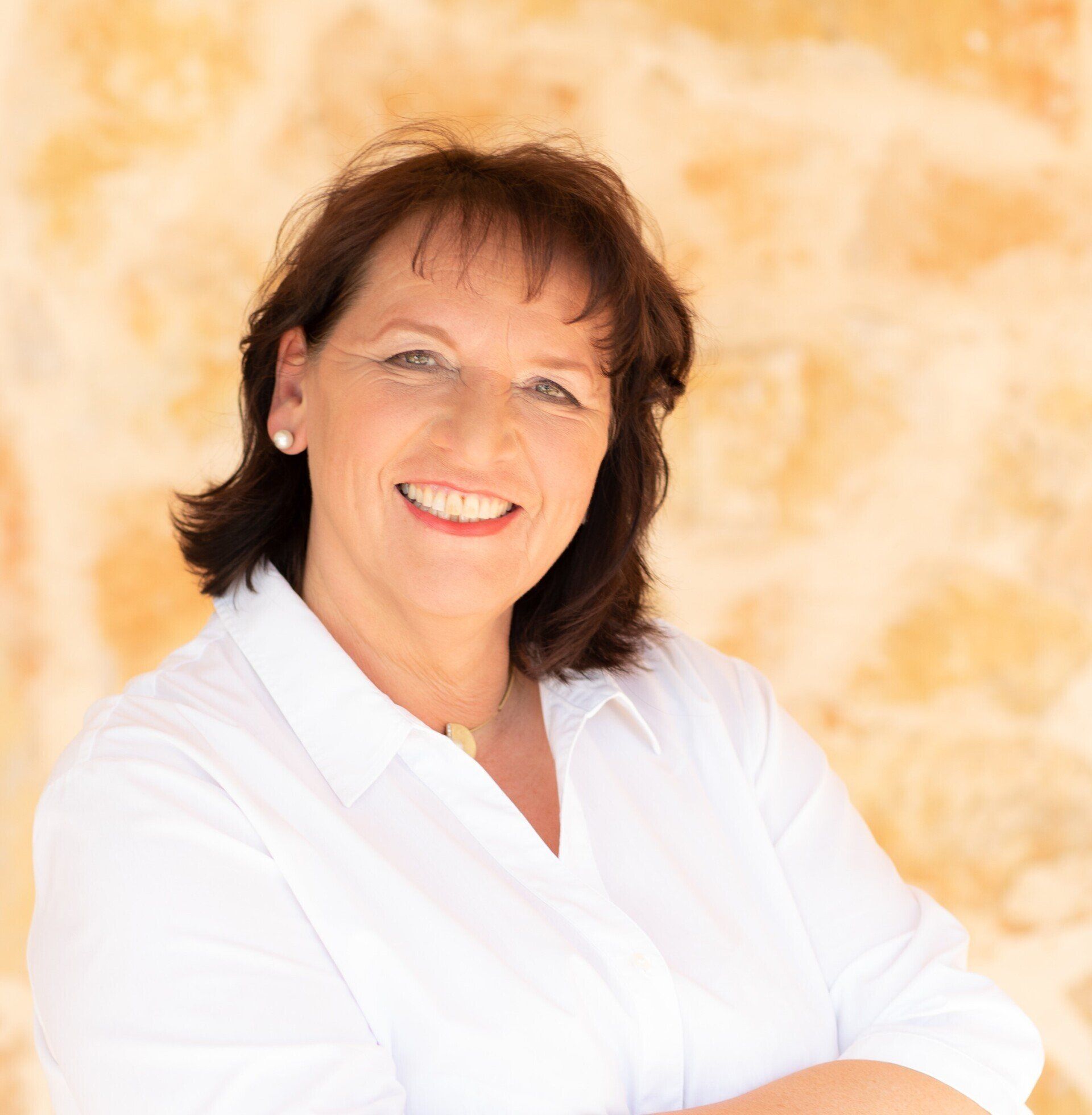 Mujer de cabello oscuro sonríe, vistiendo una camisa blanca; fondo de pared de piedra.