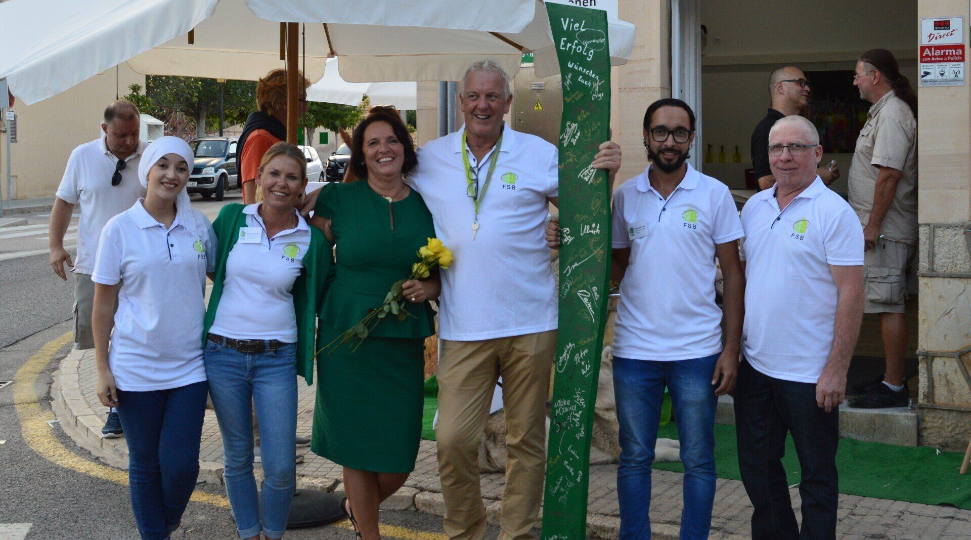 Grupo de seis personas sonriendo, sosteniendo un cartel verde al aire libre frente a un edificio.