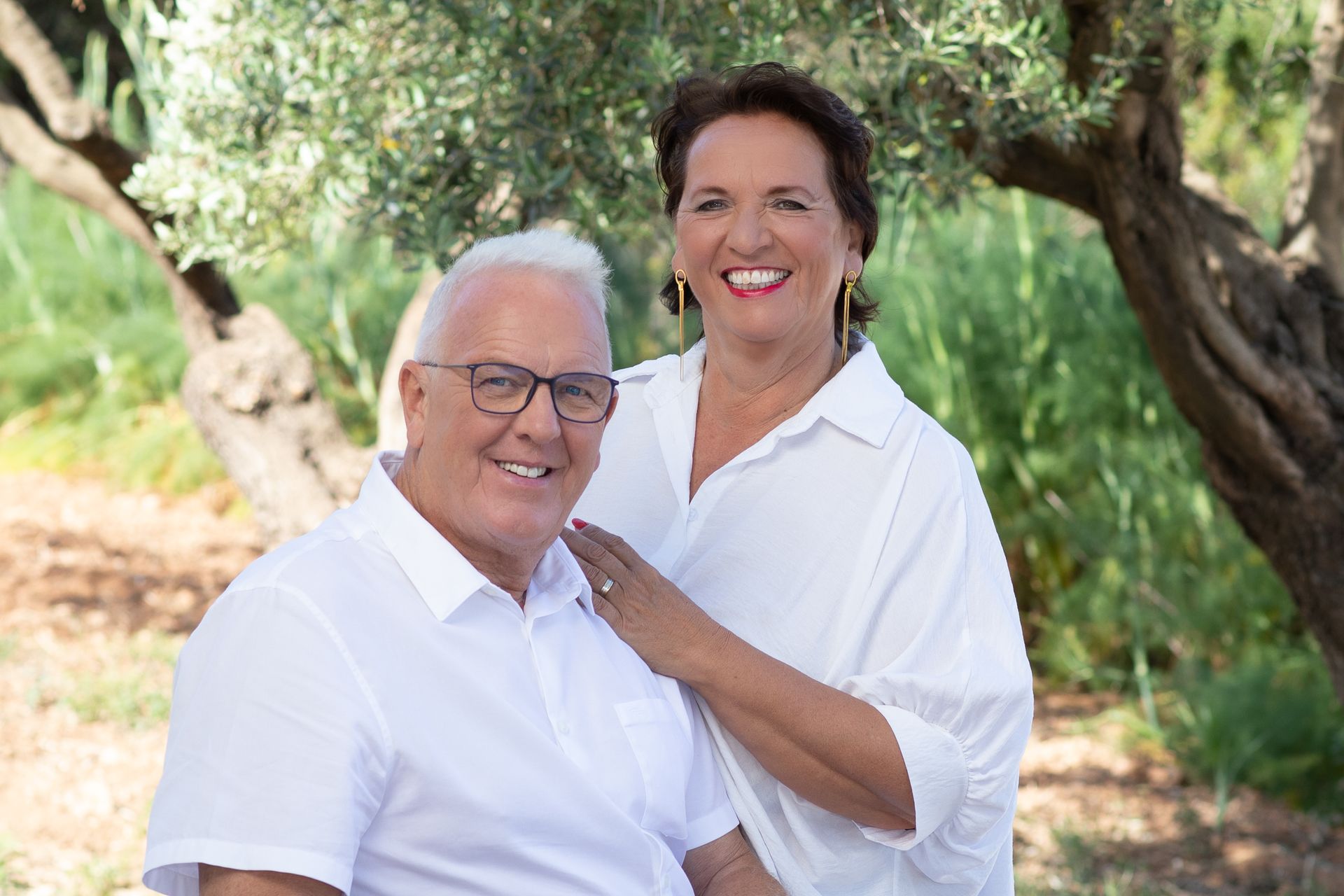 Pareja sonriendo con camisas blancas al aire libre, mano de mujer en el hombro del hombre, olivo de fondo.