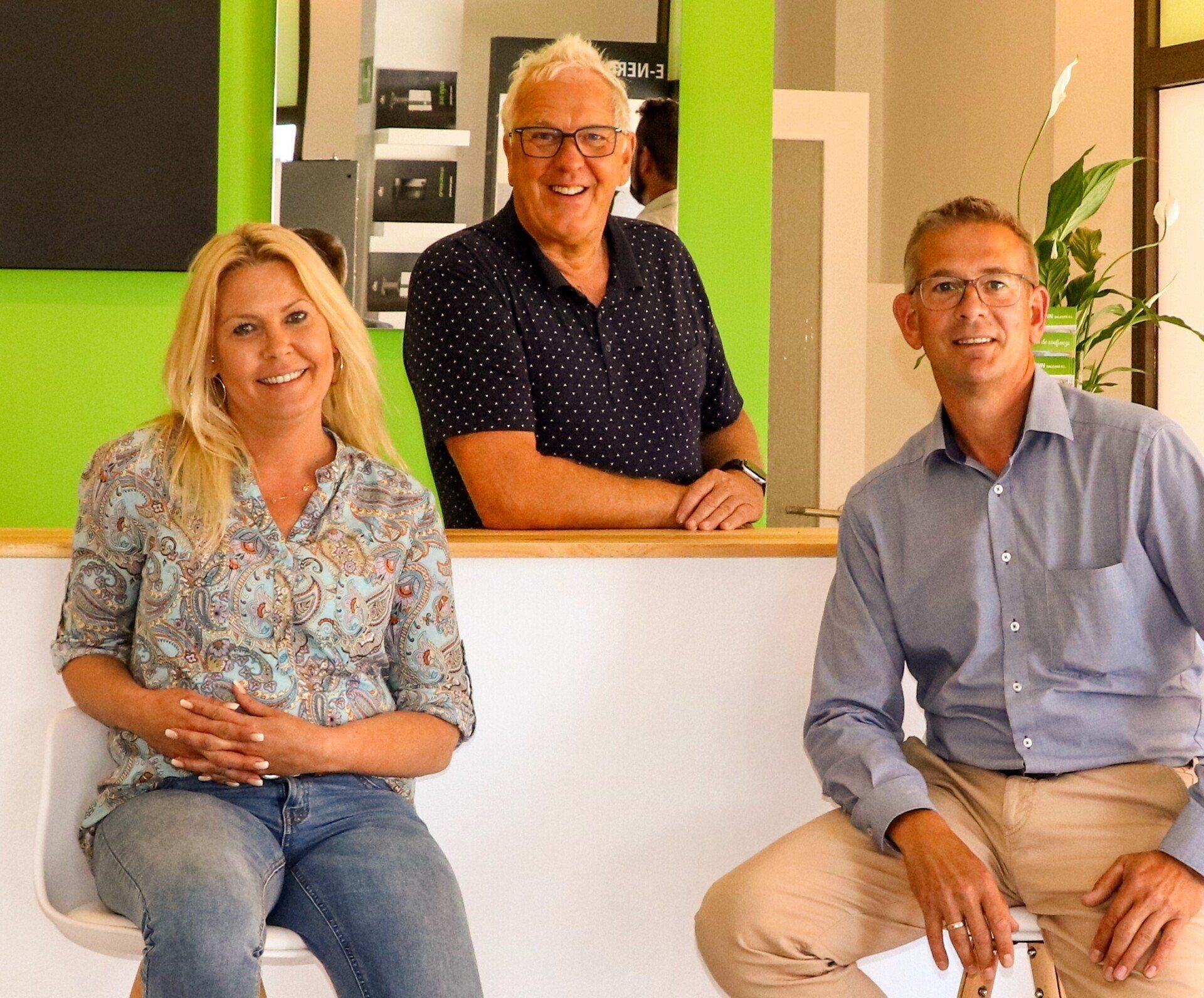 Three people smiling, posed behind a counter. Green wall in background.