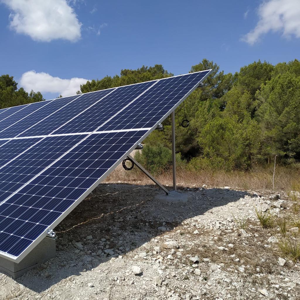 Solar panels angled toward the sky, set in a gravelly field, with trees in the background under a blue sky.