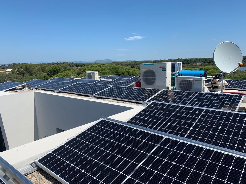Paneles solares instalados en un tejado blanco bajo un cielo azul, con unidades de aire acondicionado y una antena parabólica visibles.