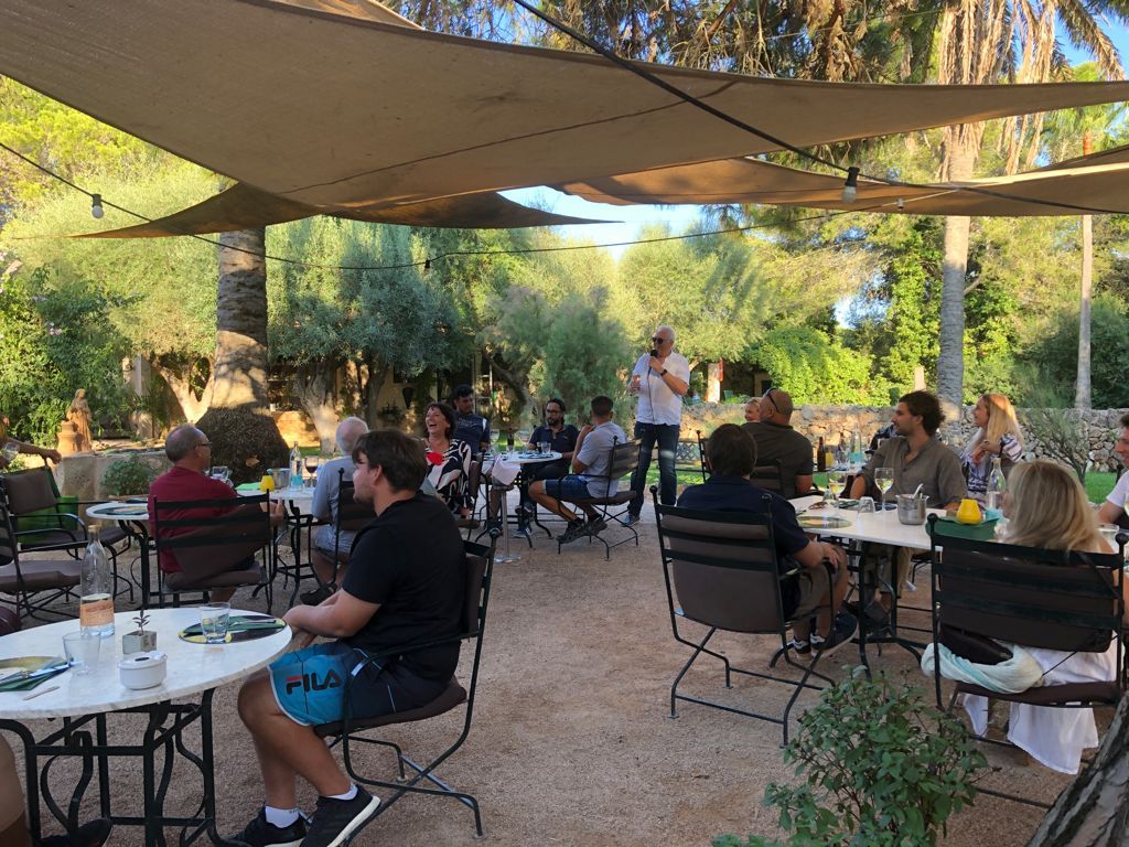 Grupo de personas en una cafetería al aire libre, a la sombra de un dosel, escuchando a un orador. Mesas y sillas sobre grava.