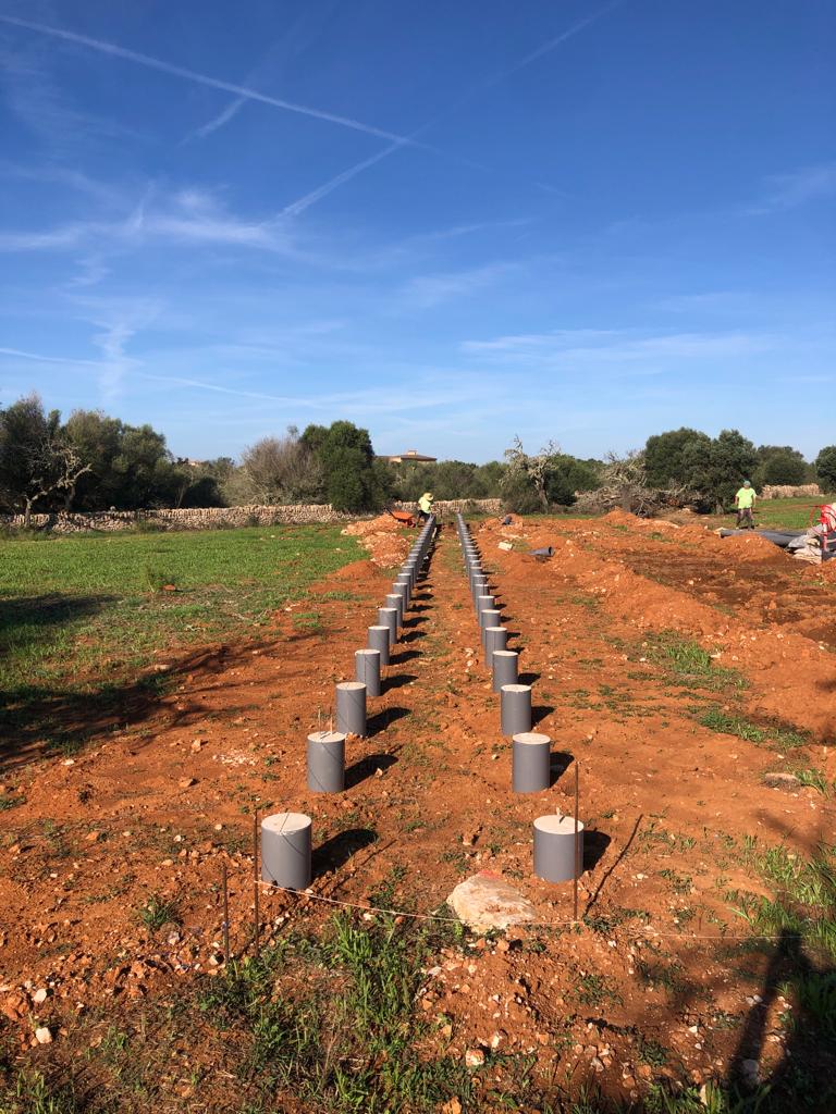 Hileras de pilares de hormigón en un campo, en una obra de construcción. Dos trabajadores al fondo, con el cielo azul por encima.