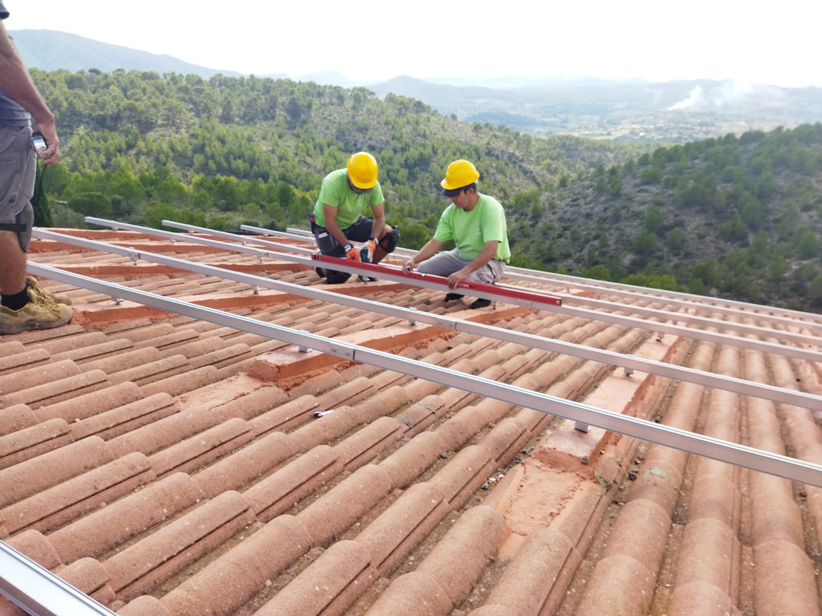 Trabajadores con cascos instalando rieles para paneles solares en un techo de tejas, con fondo montañoso.
