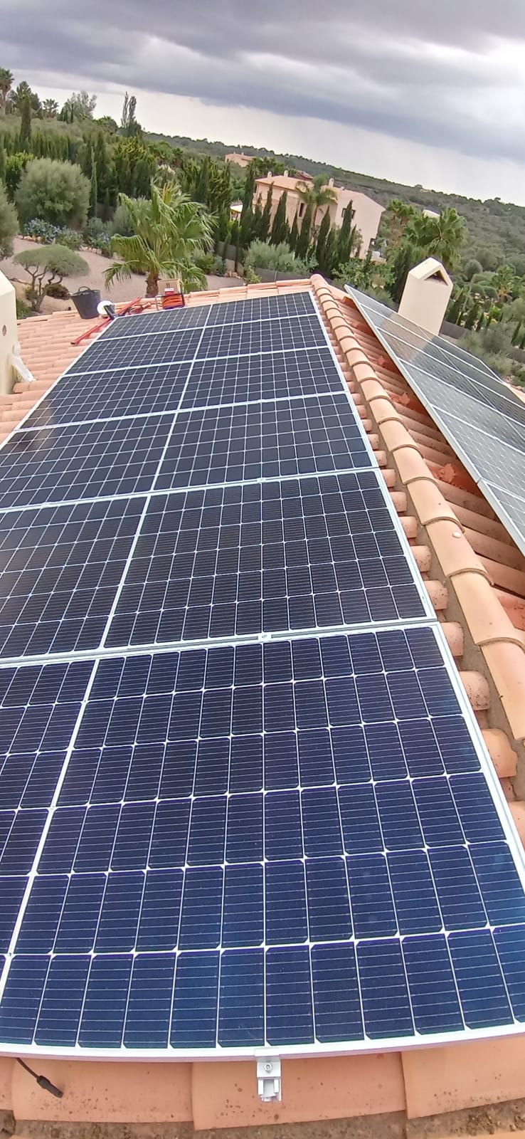 Paneles solares instalados en un techo de tejas rojas, con vista a un cielo nublado y una ladera verde.